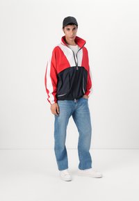 Rød, hvit og marineblå vindjakke med glidelås, elastiske ermer og to frontlommer; kombinert med lyseblå jeans og hvite joggesko.
