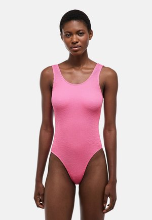 Femme mince aux cheveux courts portant un body rose texturé sans manches, debout devant un fond clair uni.