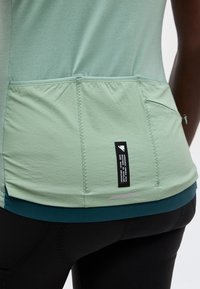 Haut de sport vert clair avec trois poches arrière et une étiquette noire, porté sur un bas de sport bleu sarcelle et noir.