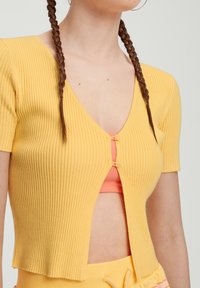 Gelbes, geripptes Kurzarmoberteil mit tiefem V-Ausschnitt und zwei Knöpfen vorne. Über einem pfirsichfarbenen Unterkleid getragen.