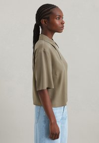 Olivgrünes Kurzarm-Bluse mit Knopfverschluss und Kragen; weicher Stoff und lockere Passform; kombiniert mit hellblauen Jeans.