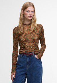 Langarmtop in donkergroene stof met een oranje bloemenpatroon, voorzien van een ronde hals en een getailleerd ontwerp, gecombineerd met high-waisted blauwe jeans.