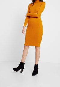 Ribbed turtleneck w żółtym musztardzie, dopasowany, o długości do kolan. W połączeniu z czarnymi zamszowymi botkami za kostkę, z detalem złotego zamka.