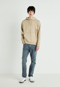 Sudadera de algodón beige con un bolsillo frontal y capucha ajustable, combinada con unos vaqueros de mezclilla azul claro y zapatillas blancas con detalles en marrón.