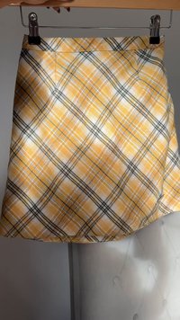 Gonna a quadri gialli e bianchi con linee nere, appesa a una gruccia di metallo su uno sfondo semplice.