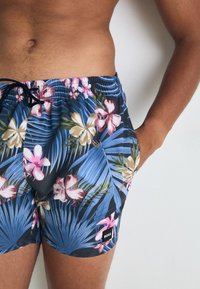 Torso masculin îmbrăcat cu șorturi de baie florale albastre, cu flori roz și bej, frunze verzi și un șnur negru, având mâna în buzunar, pe fundal alb.