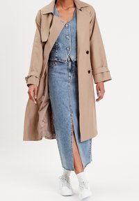Trench-coat beige porté sur un gilet court en jean et une jupe taille haute, avec une fente à l'avant, des ourlets usés, et des baskets blanches avec des accents verts.