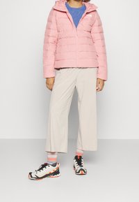 Veste rose rembourrée avec capuche, fermeture éclair à l'avant et coutures horizontales. Associée à des pantalons larges beige et des baskets multicolores.
