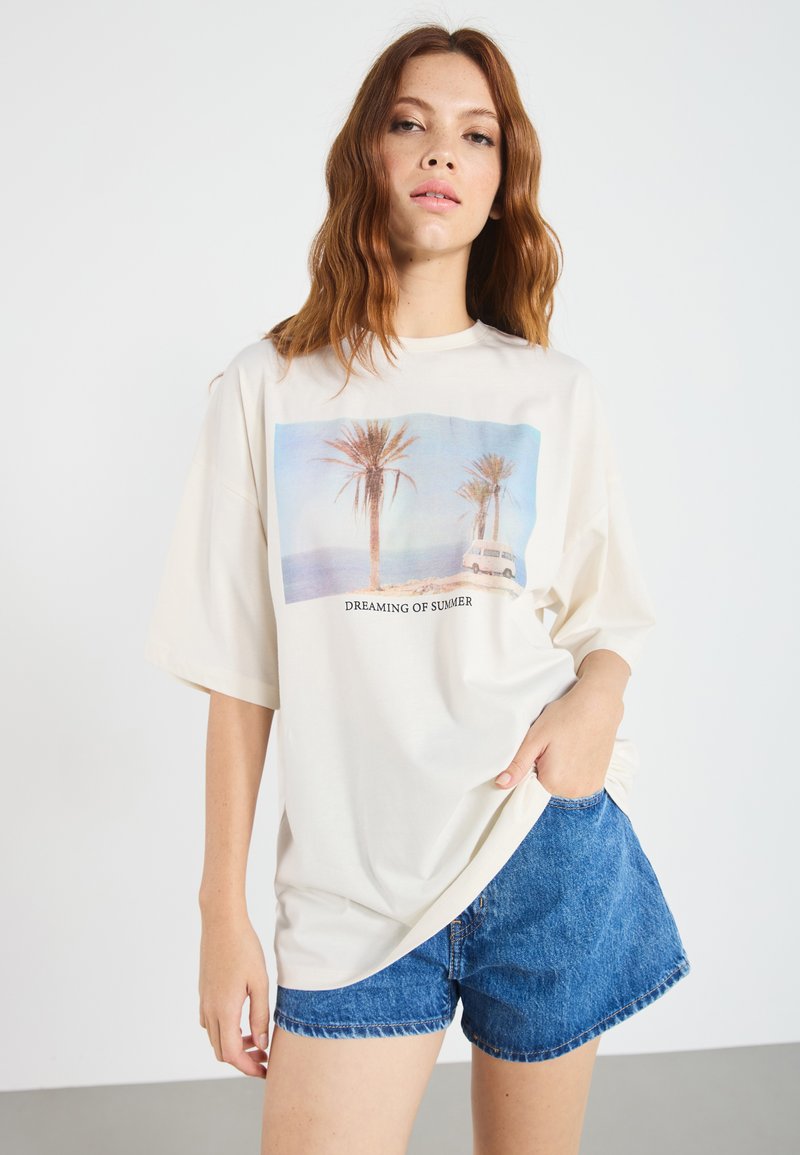 Cream oversized t-shirt with a graphic of palm trees and a vehicle, captioned "DREAMING OF SUMMER." Soft fabric, relaxed fit, short sleeves.