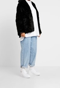 Chaqueta de piel sintética negra, diseño oversized, llevada sobre una camisa blanca de botones y unos vaqueros holgados de color azul claro, combinada con zapatillas blancas.