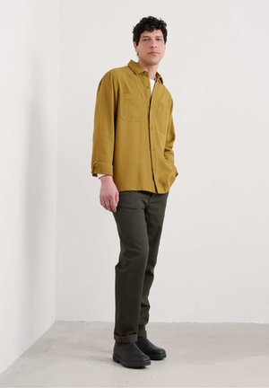 Mosterdgele blouse met lange mouwen en twee voorkwasten, gecombineerd met donkergroene broek en zwarte instappers, tegen een neutrale achtergrond.