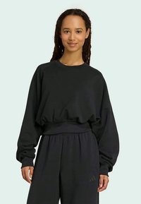 Melnas oversized džemperis ar garajām piedurknēm, apaļu kakla izgriezumu un ribotu apakšmalu; audums izskatās mīksts un struktūrēts.