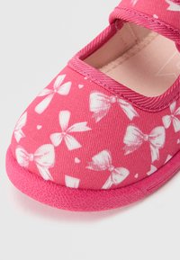 Pantof roz din material textil, cu un design modelat format din funde și inimioare albe; prezintă un vârf rotunjit, o brățară elastică și o talpă texturată roz.