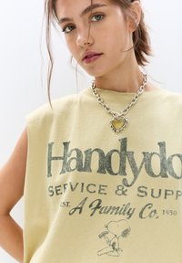 Gul ærmeløs skjorte med grafisk tryk, der står "Handydots Service & Support" med Snoopy. Kraftig sølv kædehalskæde med hjerte vedhæng.