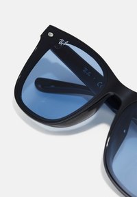 Ochelari de soare din plastic negru cu lentile albastre, având o formă de ochi de pisică, accent metalic și logo-ul Ray-Ban pe ramă și lentilă.