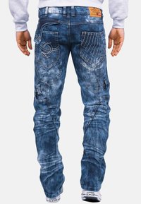 Jeans in denim blu con un aspetto sbiadito e rovinato, dettagliati da cuciture bianche, numerose tasche e un'etichetta in pelle sulla vita.