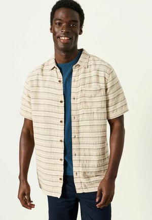 Lächelnder Mann, der ein beigefarbenes gestreiftes Kurzarmhemd über einem blauen T-Shirt mit Rundhalsausschnitt und dunkler Hose trägt, vor einem einfarbigen Hintergrund stehend.