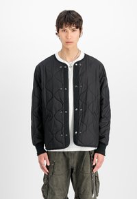 Chaqueta acolchada negra con cuello redondo, cierre de botones a presión, puños acanalados y patrón texturizado, combinada con pantalones cargo verde oliva.