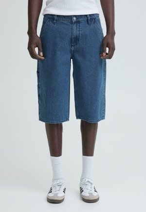 Mand iført blå denimshorts til knæene, hvide sokker, hvide og grå sneakers og en hvid ærmeløs top, stående mod en ensfarvet baggrund.