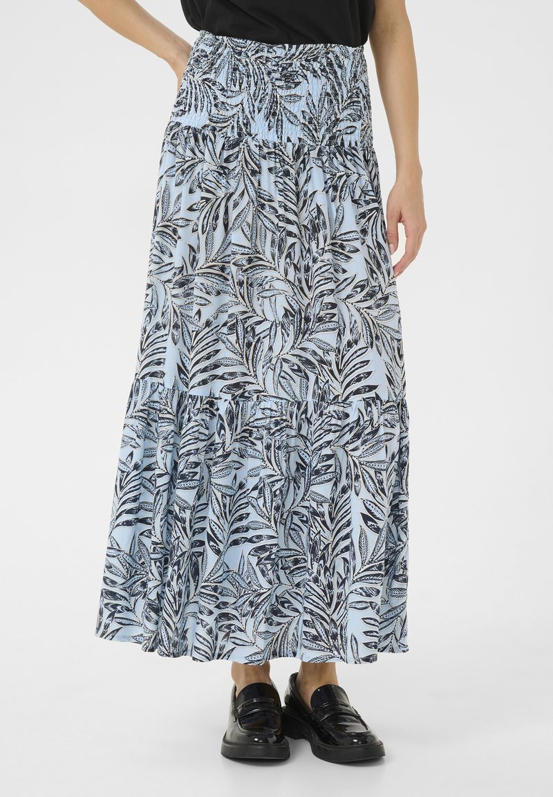 Jupe maxi en tissu bleu clair avec imprimé de feuilles noires. Présente une taille smockée et un design fluide à volants. Associée à des mocassins noirs.