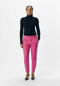 Pull noir près du corps avec un col roulé associé à un pantalon rose sur mesure, en tissu texturé, et des escarpins noirs à bout pointu. Design simple et moderne.