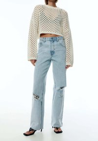 Pull court en maille crochet blanche à manches longues, associé à un jean déchiré taille haute bleu clair et des sandales noires à enfiler.