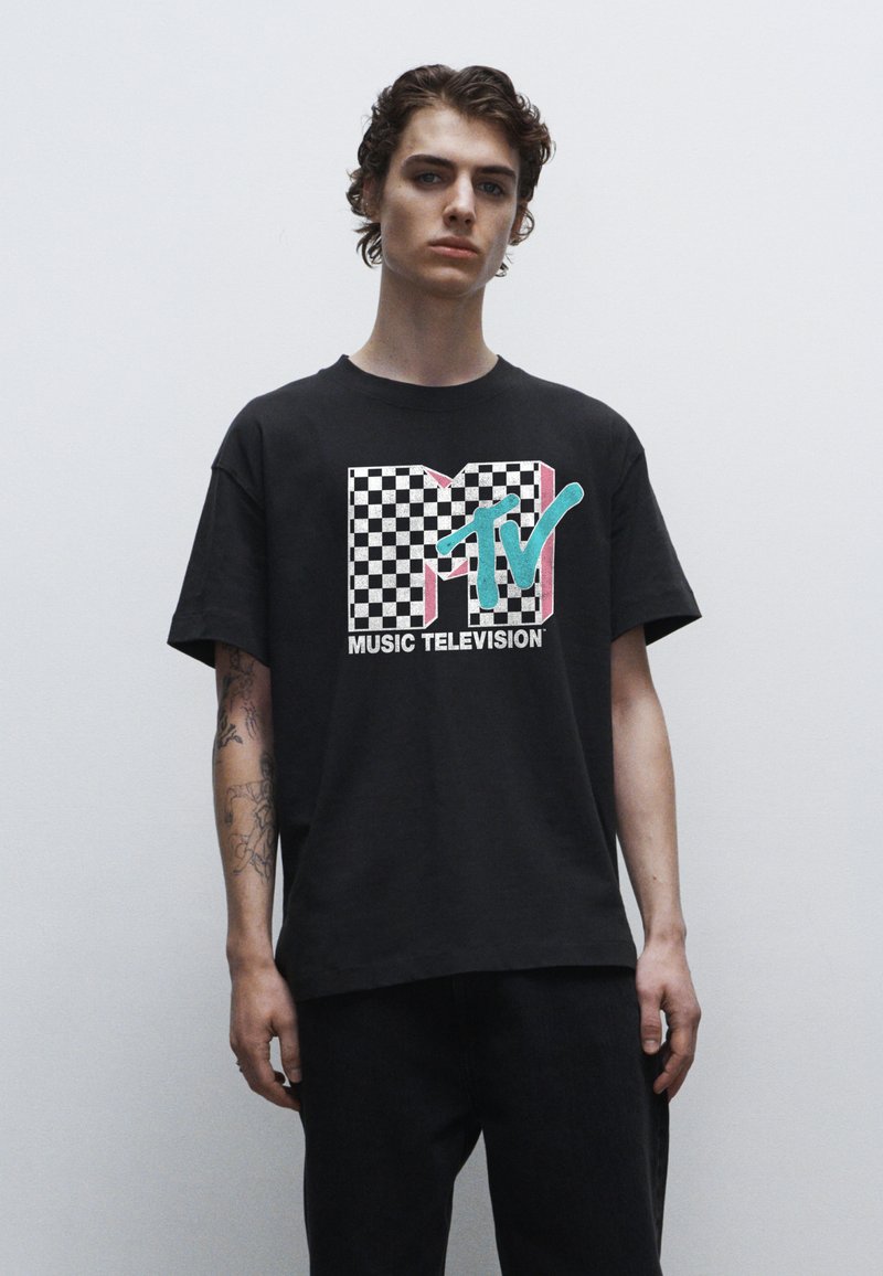 Černé bavlněné tričko s vícebarevným logem MTV se šachovnicovým vzorem. Krátké rukávy a volný střih, nošeno s tmavými džíny.
