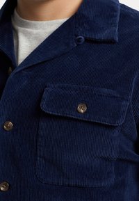 Marinblå kordjacka med en strukturerad yta, nedvikbar krage, framficka och bruna knappaccenter. Har synlig ribbning.