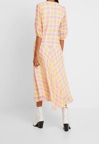 Keltainen ruudullinen mekko, jossa on puffatut hihat ja laskeutuva A-linjainen siluetti, yhdistetty valkoisiin korkokenkiin. Sujuva kangas, monivärinen ruutukuvio.