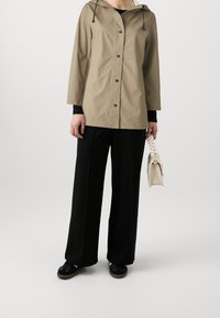Chaqueta impermeable beige con capucha, botones frontales y detalles negros, combinada con pantalones de pierna ancha negros y un bolso de color claro.