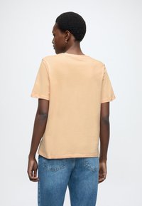 Beżowa koszulka z krótkim rękawem wykonana z miękkiej bawełny, o luźnym kroju i prostym dekolcie, zestawiona z jasnoniebieskimi jeansami.