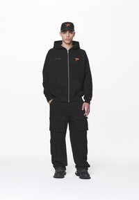 Svart hettejakke med orange logo, kombinert med svarte cargo-bukser med flere lommer. Svarte sporty sko fullfører antrekket.