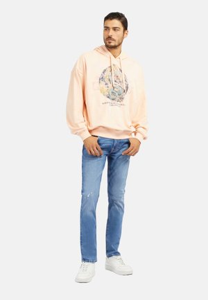 Hoodie en coton pêche avec impression graphique, coupe décontractée, poignets côtelés et poche kangourou, associé à un jean déchiré bleu clair et des baskets blanches.