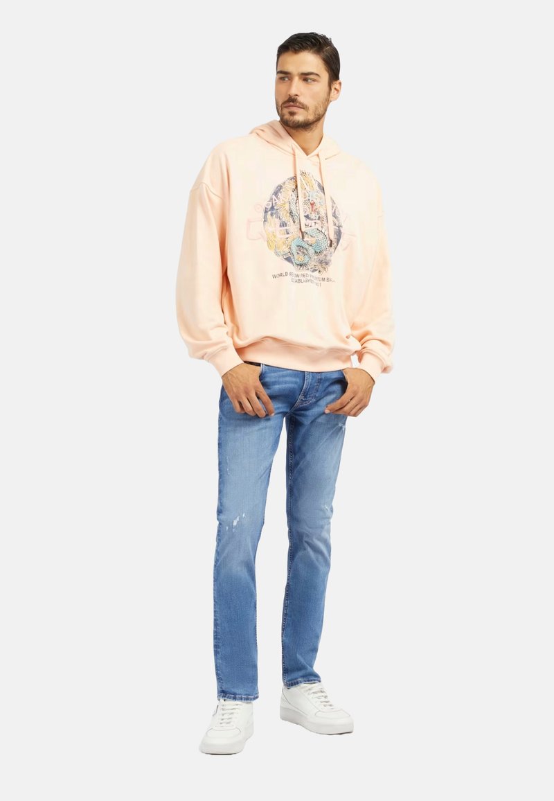 Hoodie en coton pêche avec impression graphique, coupe décontractée, poignets côtelés et poche kangourou, associé à un jean déchiré bleu clair et des baskets blanches.
