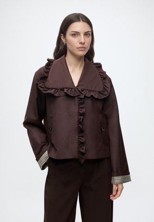 Frau trägt eine dunkelbraune Jacke mit großem Rüschenkragen und passenden Hosen, stehend vor einem weißen Hintergrund.