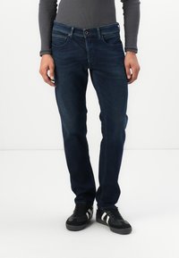Vaqueros de mezclilla de color azul oscuro con un corte recto, diseño de cinco bolsillos y cierre de botón marrón. Se llevan con zapatillas deportivas negras.