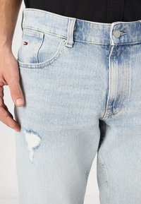 Jeans de mezclilla azul claro con un aspecto deslavado, que presentan un desgarrón en el muslo y una pequeña etiqueta de logo en el bolsillo trasero. Diseño clásico de cinco bolsillos.