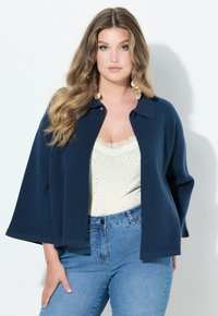 Marineblau, leichter Cardigan mit Kragen, offener Vorderseite und weiten Ärmeln, über einem cremefarbenen Tanktop mit Spitzenbesatz und Perlenakzenten getragen.