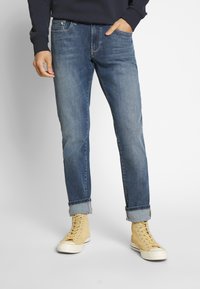 Jean en denim bleu pour homme avec une coupe droite, un léger effet délavé et des ourlets roulés, associés à des baskets beiges montantes.