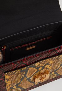 Bolso estructurado con un patrón de serpiente rojo y amarillo, que cuenta con un cierre dorado y un interior de tela negra con un bolsillo con cremallera.
