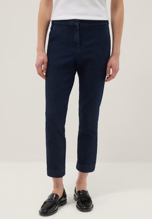 Personne portant un pantalon court bleu foncé, un haut blanc et des mocassins noirs, se tenant sur un fond clair uni.