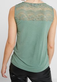 Haut sans manches en vert doux, avec une superposition en dentelle sur les épaules. Texture douce avec une coupe décontractée et un ourlet arrondi.