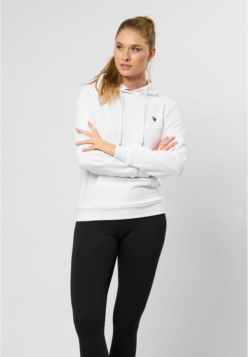 Hvit hettegenser med snøring, med en liten svart logo på brystet. Kombinert med svarte leggings, mot en nøytral bakgrunn.