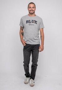 T-shirt en coton gris avec "BLUE Jeans" imprimé en bleu marine et motif à carreaux. Associé à un pantalon en denim foncé et des baskets blanches. Design simple, coupe décontractée.