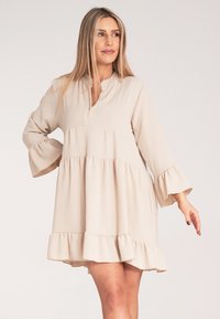 Beżowa sukienka z długim rękawem, okrągłym kołnierzykiem, falbaną u dołu i teksturowanym materiałem. Posiada sylwetkę typu A i rozkloszowane rękawy.