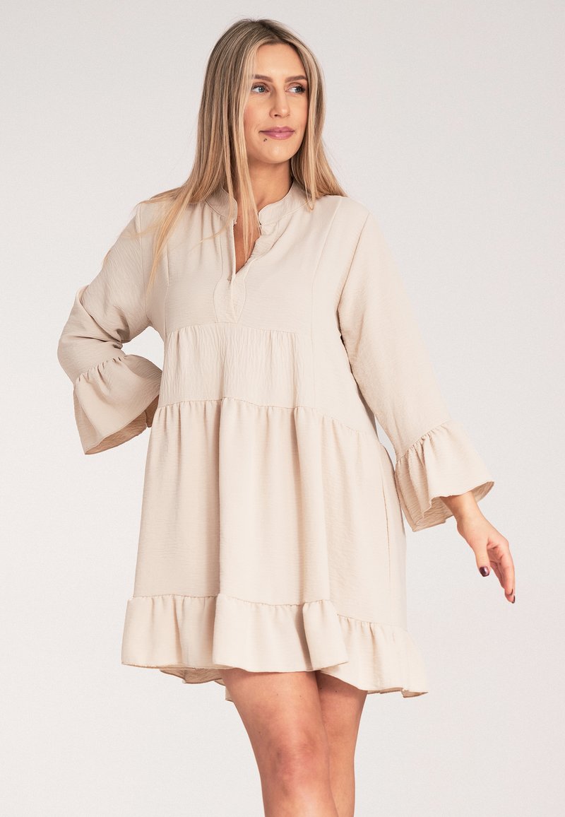 Beżowa sukienka z długim rękawem, okrągłym kołnierzykiem, falbaną u dołu i teksturowanym materiałem. Posiada sylwetkę typu A i rozkloszowane rękawy.