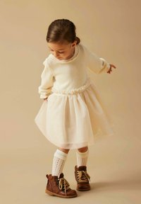 Pull en tricot crème avec taille volantée, jupe en tulle superposée, chaussettes crème montant aux genoux, et bottines marron à lacets.