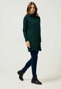 Maglione oversized verde scuro con collo a barchetta, realizzato in tessuto a maglia testurizzata, indossato con jeans skinny blu e stivaletti neri alla caviglia.