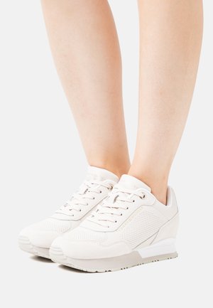 White sneakers featuring a mix of smooth and perforated materials, round toe, and a subtle wedge heel with gold branding accents.