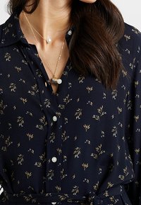 Granatowa bluzka z małym wzorem kwiatowym w odcieniach beżu i niebieskiego, z kołnierzykiem, zapięciem na guziki i paskiem w talii. Złote naszyjniki z koralikami.